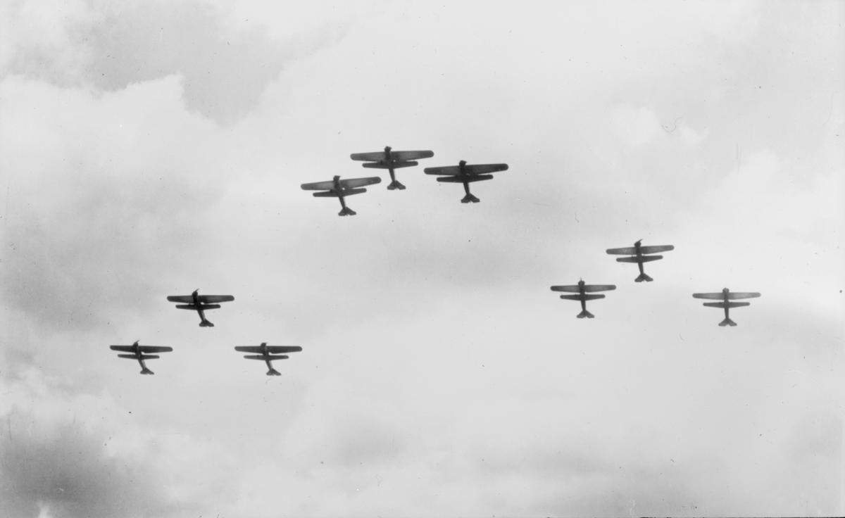 Motiv: Nio stycken flygplan flyger i V-formation. Flygplan S 6 och J 3. Vy nedifrån. Ur Gamla Malmen-samlingen. Tillhör Flygvapenmuseums bildarkiv.